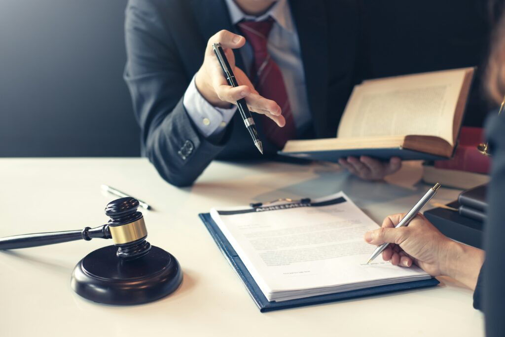 child-custody-lawyer - Pinkham & Associates, APLC A suited professional gestures with a pen while holding an open legal book, speaking across a desk where another person is signing documents on a clipboard. A judge’s gavel rests nearby, emphasizing the legal and formal setting.