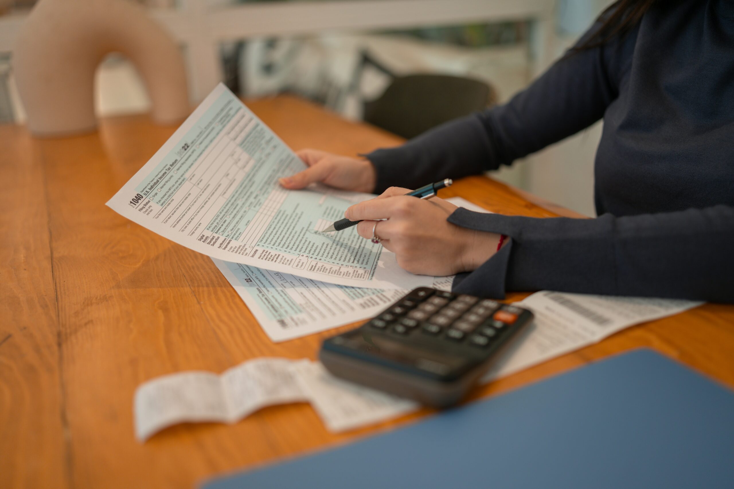 Woman sitting at a desk at home, focused on filling out tax forms with papers and a calculator nearby. - Pinkham & Associates, APLC Woman sitting at a desk at home, focused on filling out tax forms with papers and a calculator nearby.